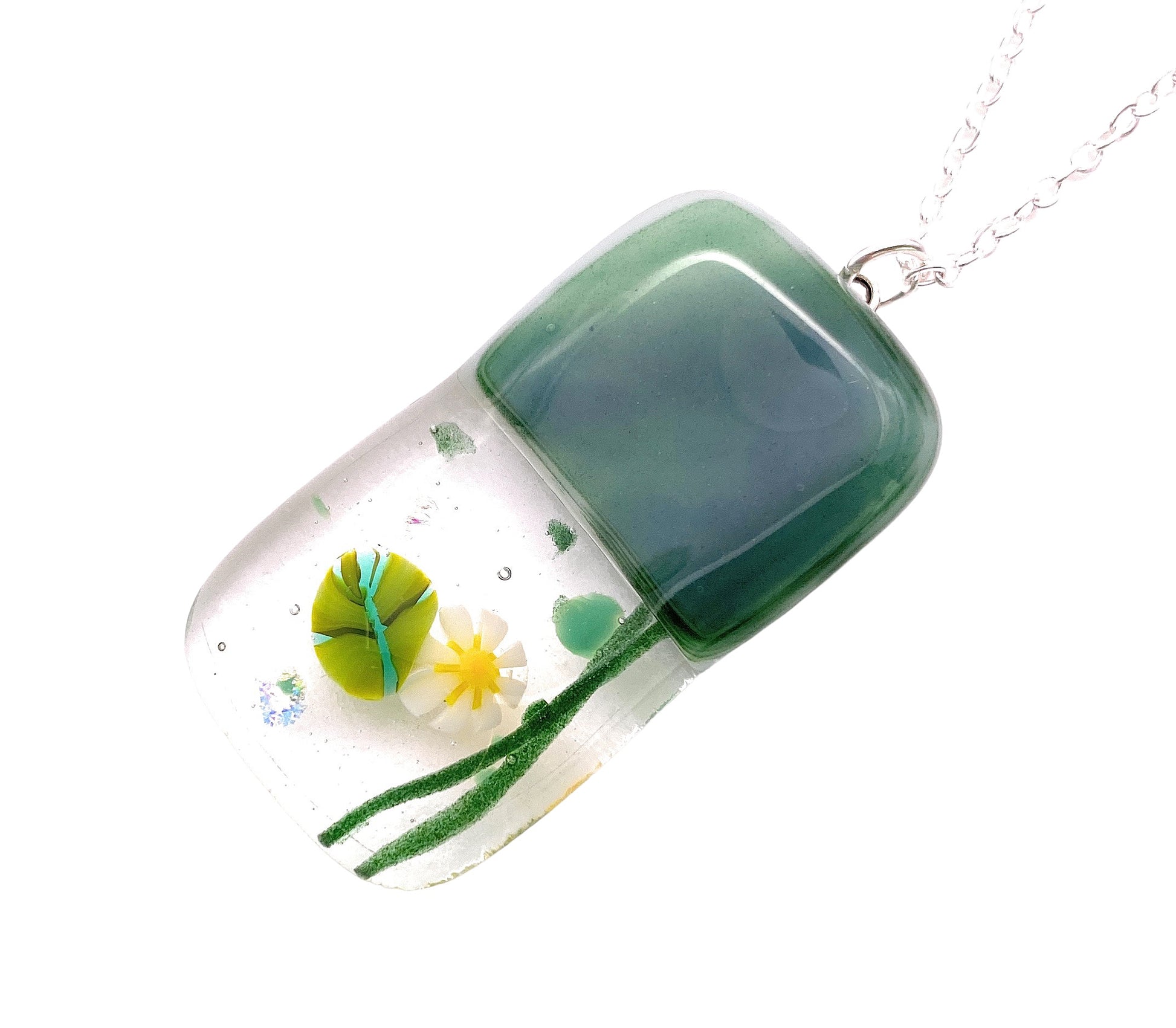 Introducing the Handmade Dichroic Glass Pendant by Calon Glass, featuring a green, rounded rectangular glass piece on a silver chain. The pendant showcases delicate green and white botanical designs, including a flower, reminiscent of millefiori glass art, with the top half adorned in solid green. Measuring 4cm by 2cm, this unique piece captures the essence of artistic elegance.