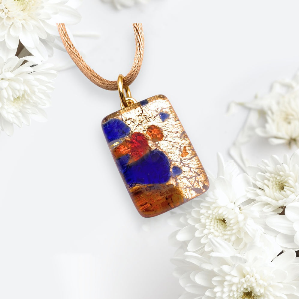 A Murano Passion Millefiori Pendant on Gold Leaf (2cm x 1.5cm) hangs from a rose-colored cord, resting on a white surface with delicate white flowers. Featuring vibrant gold, blue, red, and orange hues, it embodies elegance and comes with a Certificate of Authenticity.