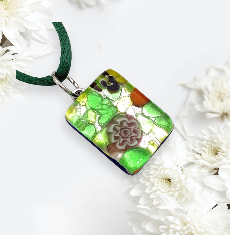 A stunning Murano Glass Pendant, named "Millefiori Flowers on Silver Leaf," in vibrant shades of green, orange, and purple from Murano Passion hangs from a dark green cord. This dainty pendant, measuring 2cm x 1.5cm (MGPP17), is elegantly surrounded by white chrysanthemums on a light background.