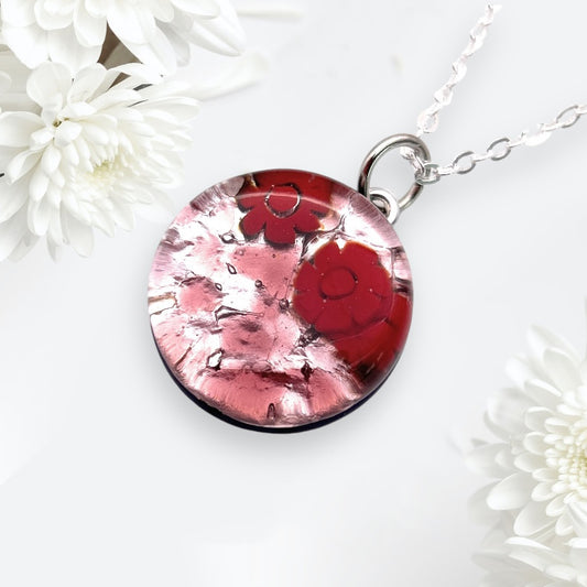 The Murano Glass Round Pendant made with Millefiori Flowers (MGPV19) by Murano Passion features a red floral design on a silver chain, complemented by white flowers. The glossy finish and intricate details highlight its exquisite craftsmanship, making it a standout piece of jewelry from the Murano Passion collection.