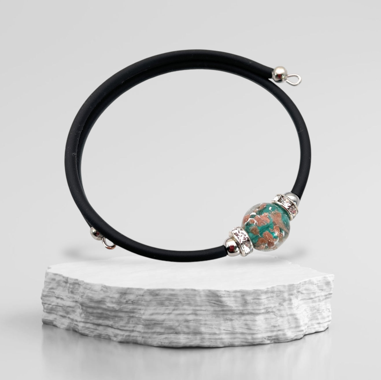 A sleek Murano Passion bracelet, featuring a single marbled 1cm Murano Glass bead on a minimalist band, is elegantly showcased on a textured white stone surface against a neutral backdrop.