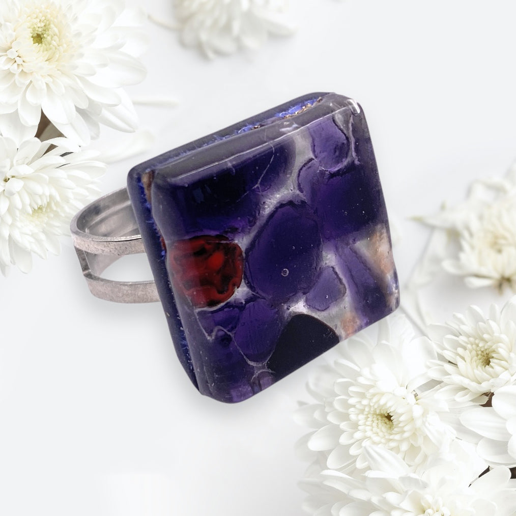 The Murano Passion Murano Glass Ring (MGR1) features a 2cm x 2cm square deep purple agate stone with unique patterns and a small reddish section, showcasing Venetian artistic heritage. Handcrafted in Italy, it's elegantly set against white daisies.