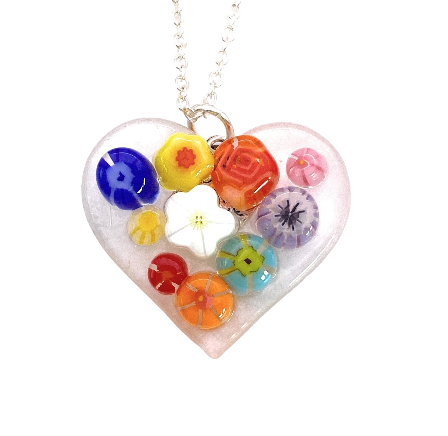 Calon Glass presents the Handmade Millefiori Flower Pendant, a heart-shaped glass piece featuring colorful geometric designs and vivid hues, elegantly suspended on a silver chain. This dainty 2.5cm pendant highlights exquisite handmade artistry with intricate floral and circular motifs.