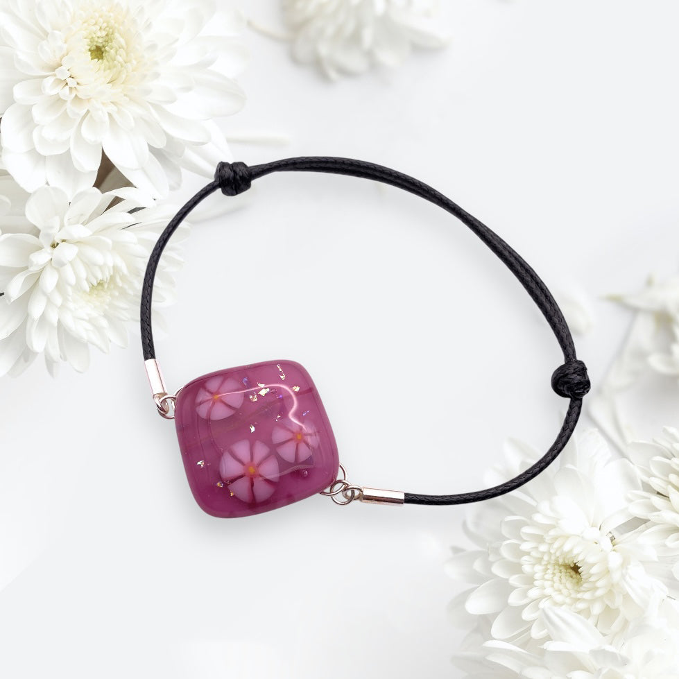 The Calon Glass Handmade Dichroic Glass Bracelet (CGBR1) features a glossy purple pendant with Millefiori flowers on a black cord, beautifully displayed among soft white chrysanthemums.