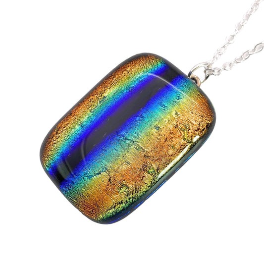 The Handmade Dichroic Glass Pendant by Calon Glass features a rectangular design embellished with vibrant stripes of blue, green, and coppery gold dichroic glass. This 3.5cm x 2.5cm kiln-fused piece is attached to a silver chain and exhibits a shimmering, iridescent effect.