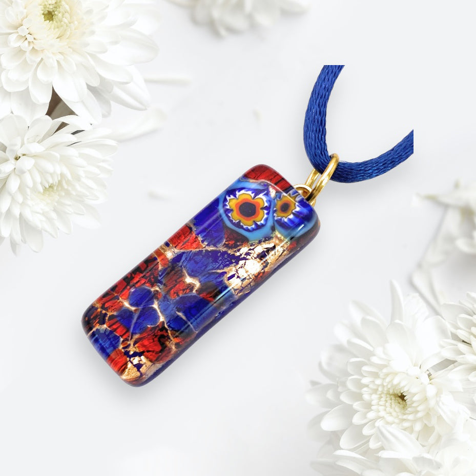 Experience the elegance of Murano Passion with this vibrant rectangular pendant, featuring intricate red, blue, and gold patterns adorned with two orange-and-blue floral designs. The pendant measures 3cm x 1cm and elegantly hangs on a blue cord amidst white flowers set against a light background. This exquisite Murano Glass Pendant with Millefiori Flowers on Gold Leaf arrives complete with a Certificate of Authenticity.