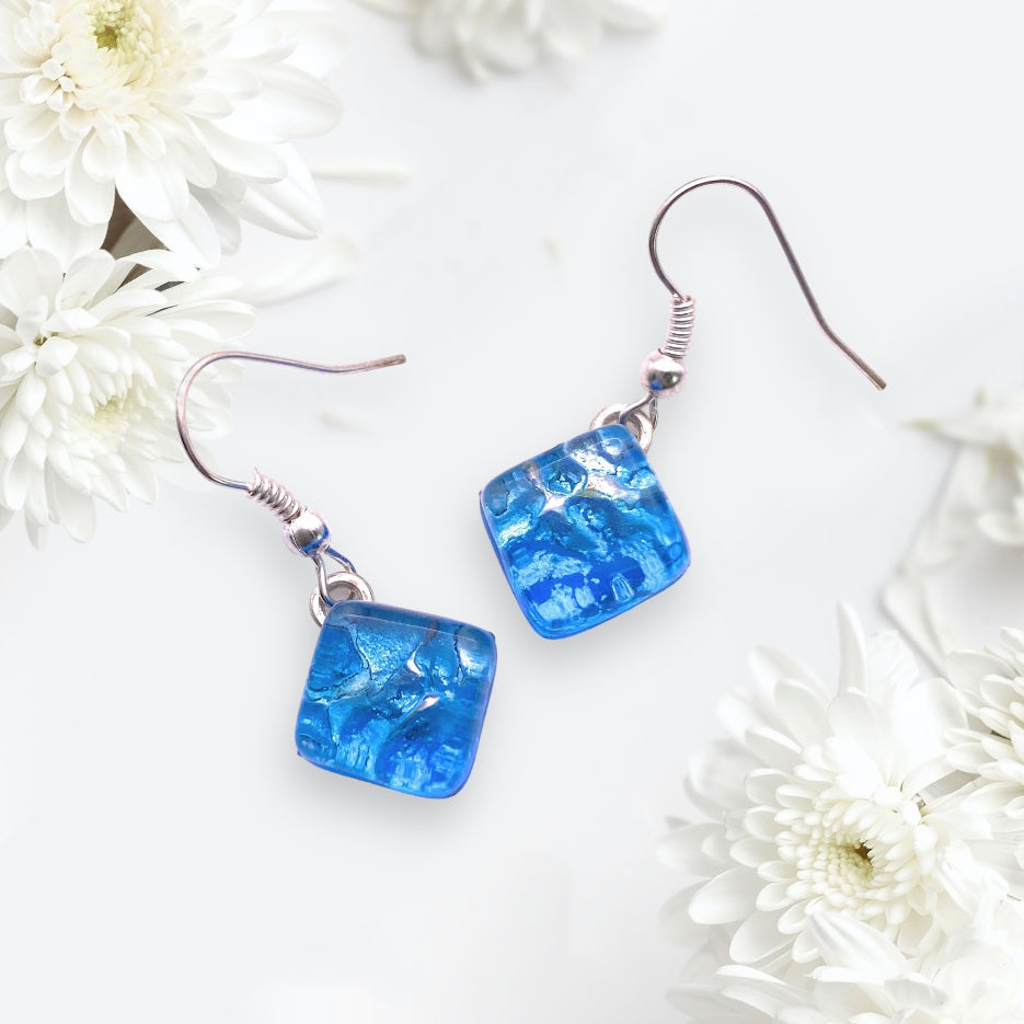 A pair of Murano Passion's Sky Blue Dropper Earrings features square-shaped Murano Glass pendants with a textured design, embodying Venetian artistic heritage. These silver wire earrings are elegantly set against a backdrop of soft white flowers on a light background.