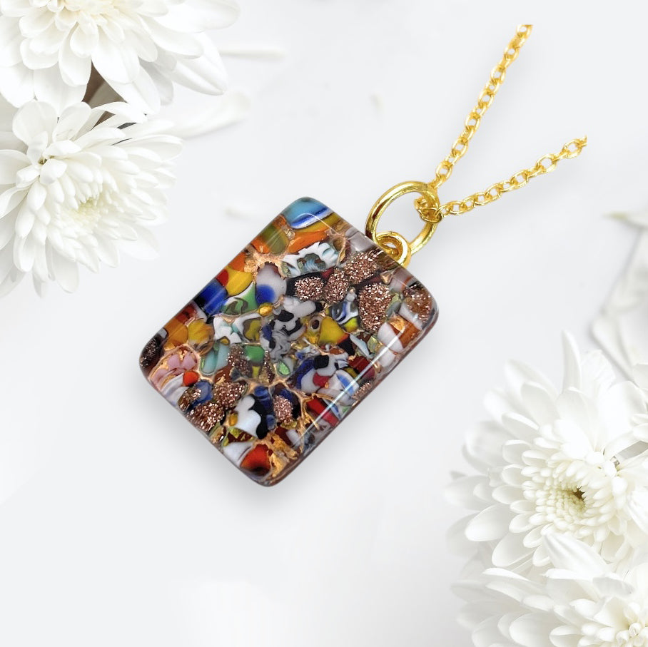 The Murano Passion Murano Glass Pendant, featuring Millefiori pieces on a gold leaf, shines gracefully among white flowers on a light background. This exquisite 2cm x 1.5cm piece showcases abstract patterns in its clear rectangular form, embodying the captivating essence of Millefiori art.