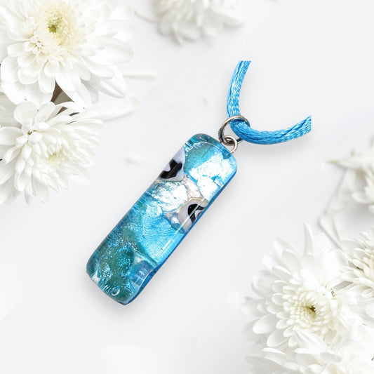 A Murano Passion Murano Glass Pendant on Silver Leaf (3cm x 1cm) sits on a matching blue cord, surrounded by white chrysanthemum flowers on a light background. With its glossy finish and abstract patterns, this pendant exudes elegance. The exquisite piece of jewelry comes with a Certificate of Authenticity.