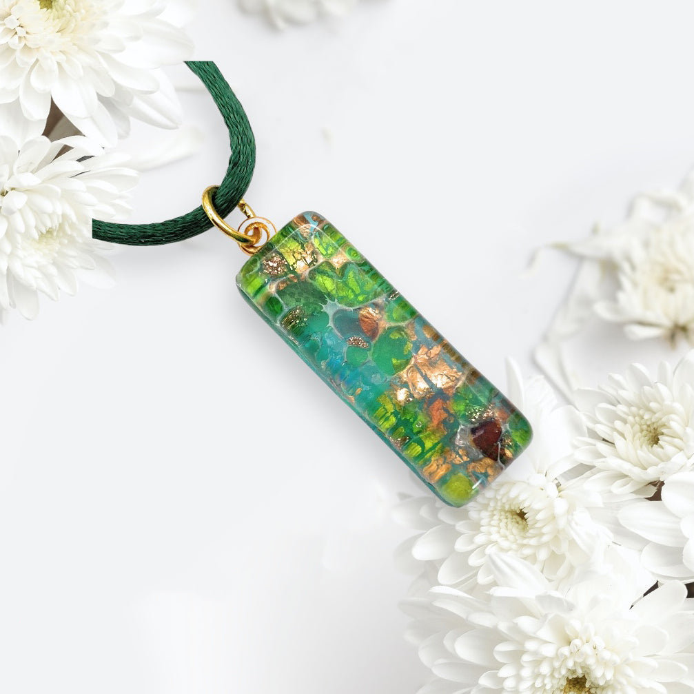 The Murano Passion product, a 3cm x 1cm rectangular pendant made with gold leaf, features a vibrant mix of green, blue, and brown hues suspended on a dark green cord. Adorned with white flowers against a light background, this Murano Glass Pendant (MGPL9) exudes the elegance of handmade jewelry artistry.