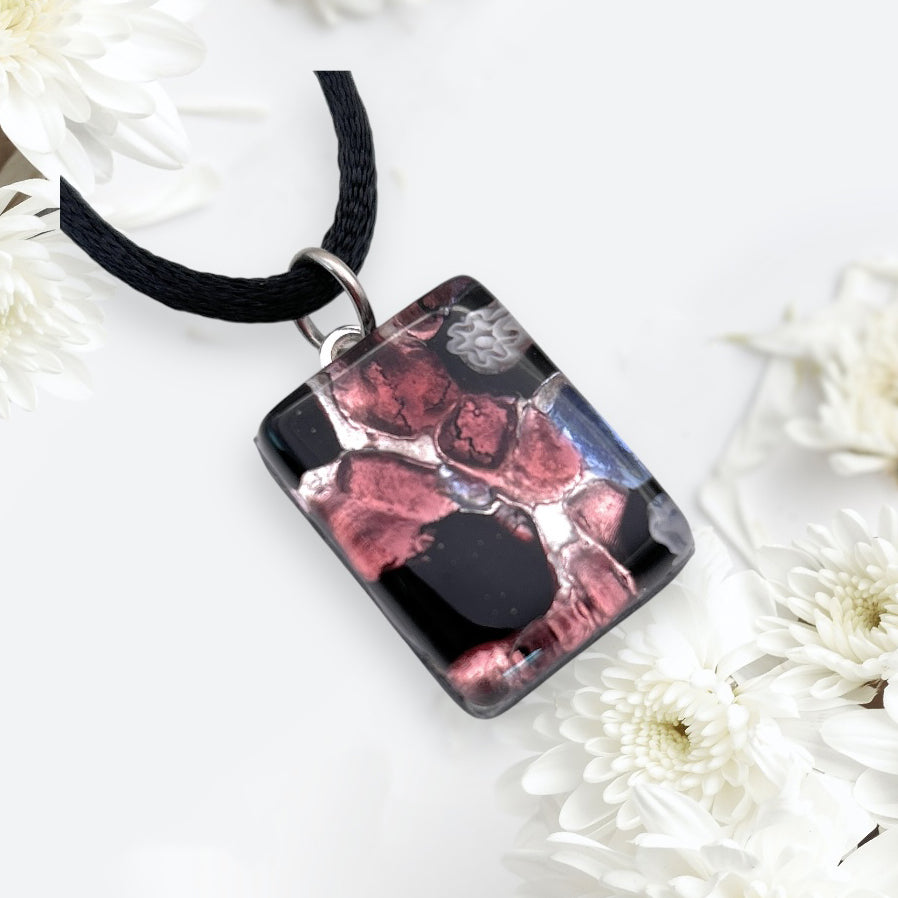 A Millefiori Flower on Silver Leaf Murano Glass Pendant from Murano Passion, measuring a dainty 2cm x 1.5cm, hangs gracefully on a black cord. It is featured against a light background adorned with delicate white flowers.