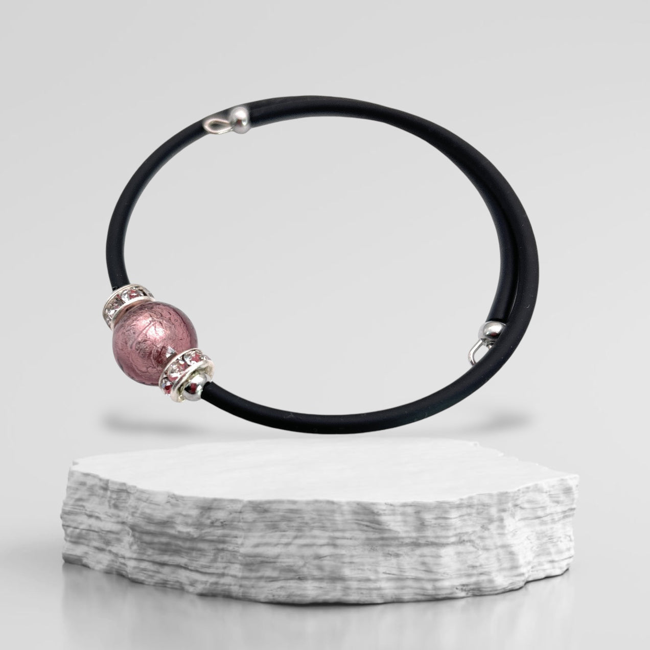 A Murano Passion black cord bracelet featuring a large 1cm pink Murano glass bead and silver accents is elegantly displayed on a textured stone platform against a plain gray background. This circular and sophisticated piece, the Murano Glass Bead Bracelet, exudes simple elegance that highlights the timeless artistry of Murano Glass Jewellery.