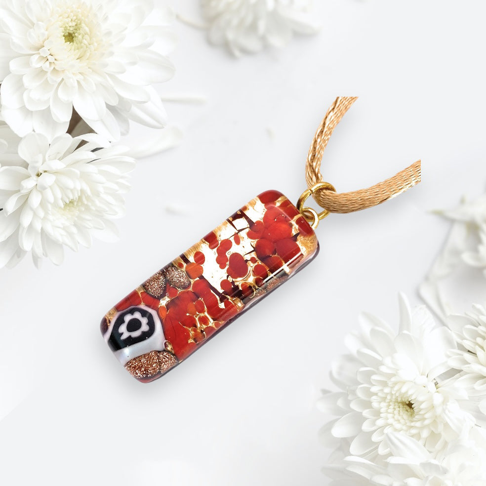 A Murano Passion MGPL12 pendant, crafted from Murano glass with a striking abstract pattern of red, black, and gold using gold leaf, measures 3cm x 1cm. It hangs elegantly from a golden chain against the soft-focus backdrop of white chrysanthemums.