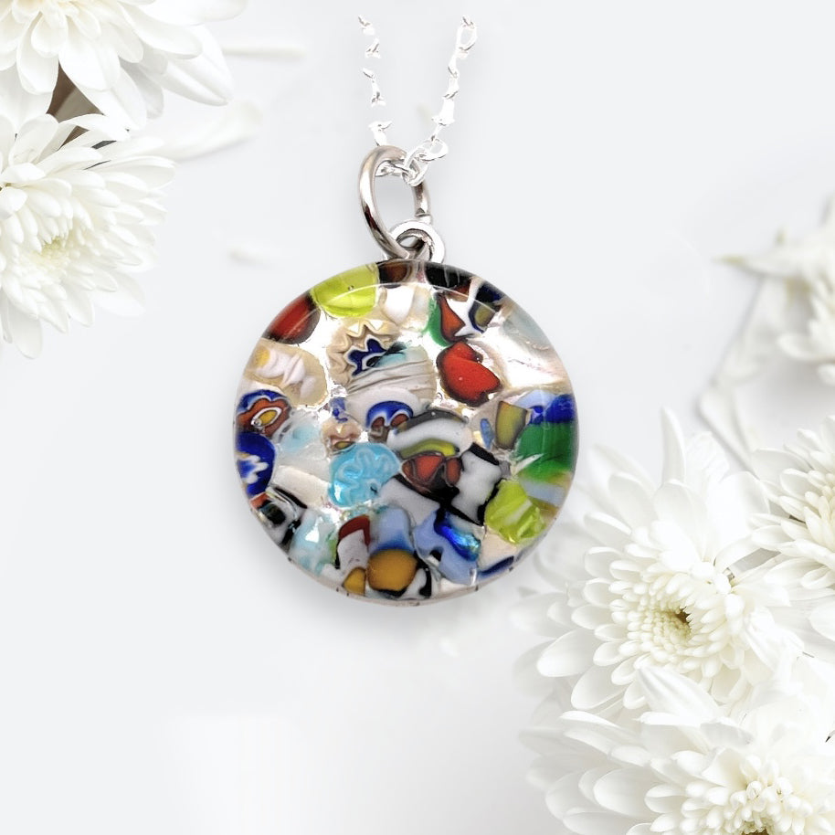 A round Murano Glass Round Pendant made with Millefiori (2cm) from the Murano Passion brand, featuring multicolored abstract designs, is elegantly suspended from a silver chain. Set against a light background and encircled by white chrysanthemums, this exquisite piece of Murano Glass Jewellery comes with a Certificate of Authenticity.