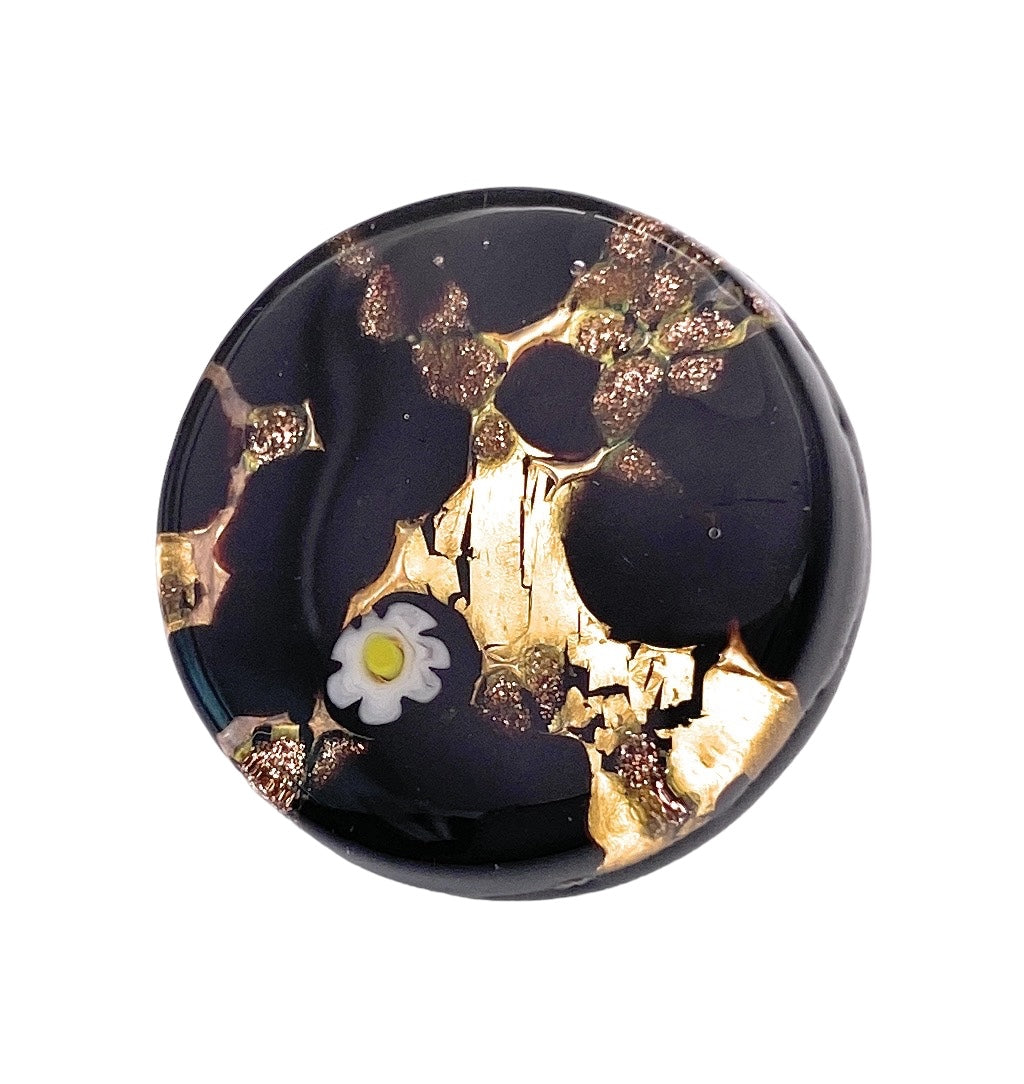 The Murano Passion brooch, named the "Murano Glass Brooch with Millefiori Flower on a Gold Leaf Base," features a captivating round glass design set against a black background, adorned with gold-colored accents and highlighted by a delicate white flower with a yellow center. This piece beautifully embodies the enchanting artistry of Murano Glass, reflecting Venetian artistic heritage through its abstract and artistic appearance.