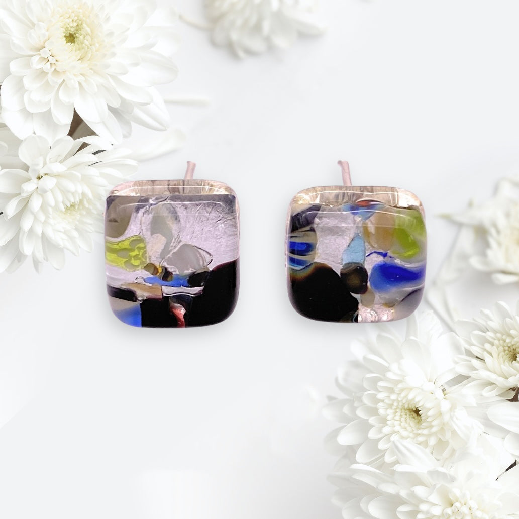 The Murano Passion earrings, product name "Murano Glass Earrings - Multi/Silver, Dainty 8mm Diameter (MGE4)," feature square Murano glass design with abstract multicolor patterns. These handmade stud earrings rest against a backdrop of white flowers and soft light, displaying shades of black, blue, green, and purple with a glossy finish that exude an artistic charm.