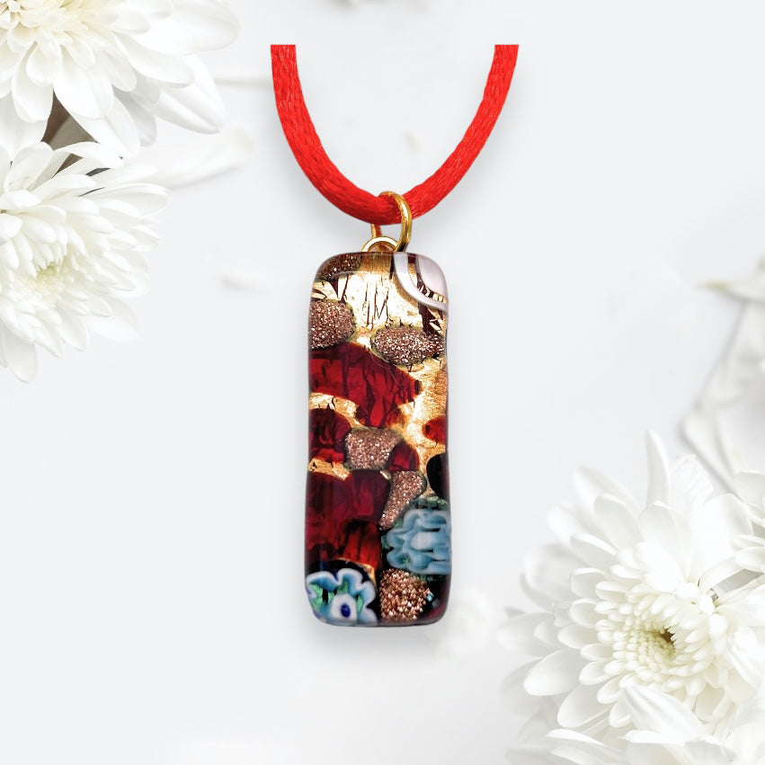 The Murano Glass Pendant by Murano Passion, made using Millefiori Flowers on a Gold Leaf, features a red cord and rectangular design adorned with intricate red, gold, and blue swirls and floral patterns. Set against a background of white flowers, this 3cm x 1cm piece (MGPL3) comes complete with a Certificate of Authenticity.