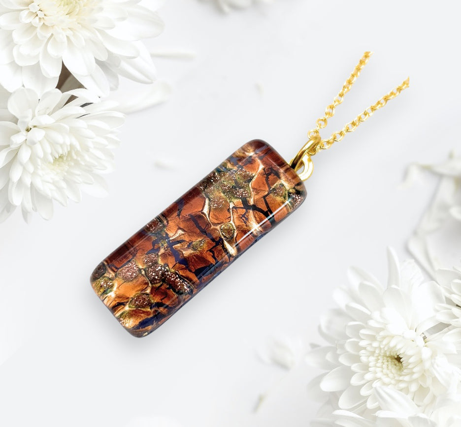 A Murano Passion pendant, crafted with gold leaf and featuring an intricate mosaic pattern in earthy tones, is showcased on a light background surrounded by delicate white flowers. This exquisite piece of Murano Glass Jewellery, measuring 3cm x 1cm (MGPL13), comes with a Certificate of Authenticity.