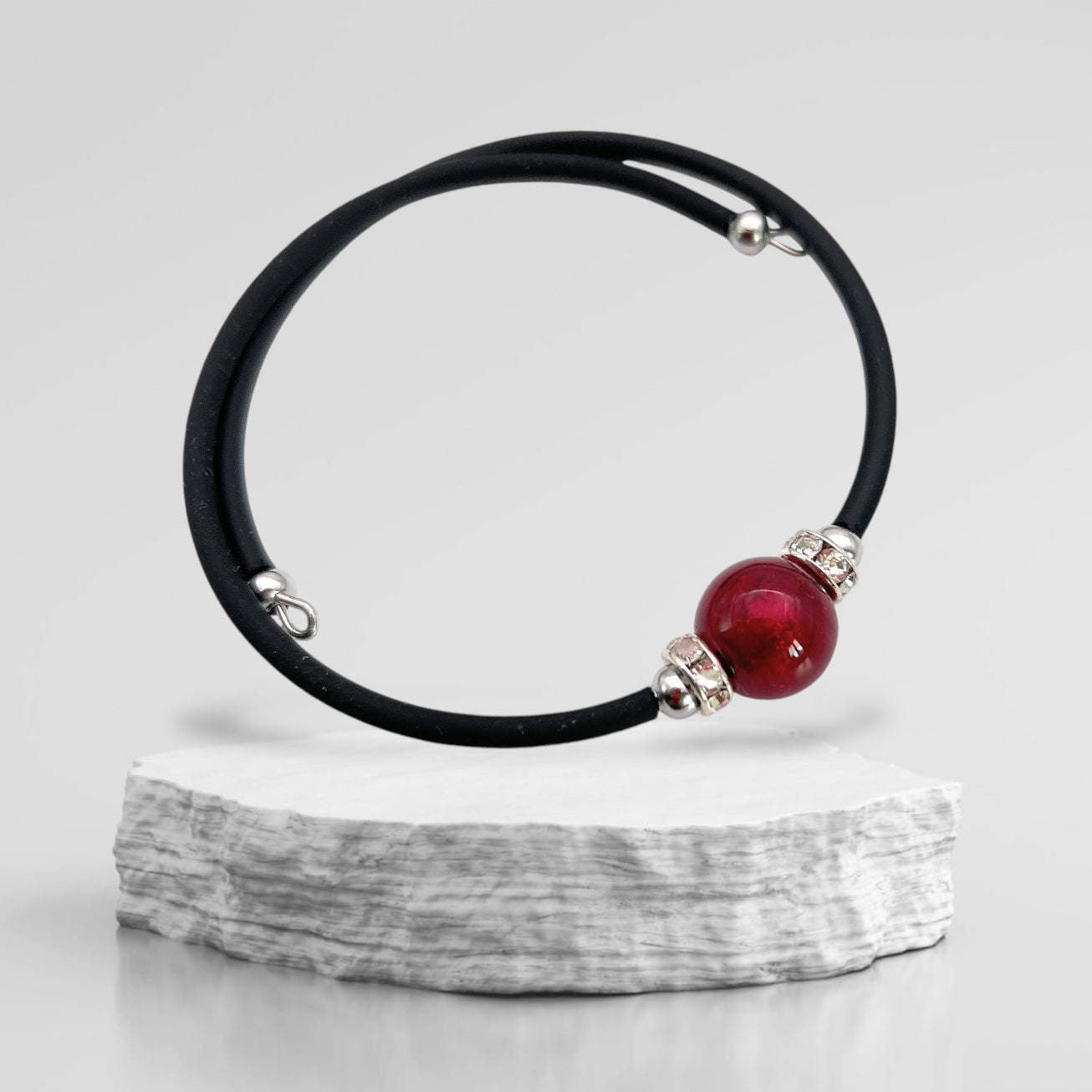 A Murano Passion bracelet featuring a genuine 1cm Murano glass bead, accentuated with silver on a sleek black cord, is gracefully showcased on a textured, round white stone platform against a gray background.