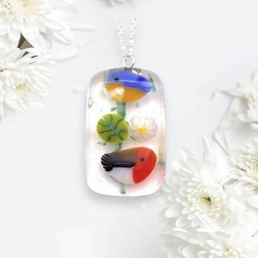 The Handmade Glass Pendant by Calon Glass (4cm x 2cm, CGP32) showcases a unique art with colorful bird and floral designs on dichroic glass set against a light background. It hangs elegantly from a delicate silver chain, epitomizing exquisite craftsmanship.