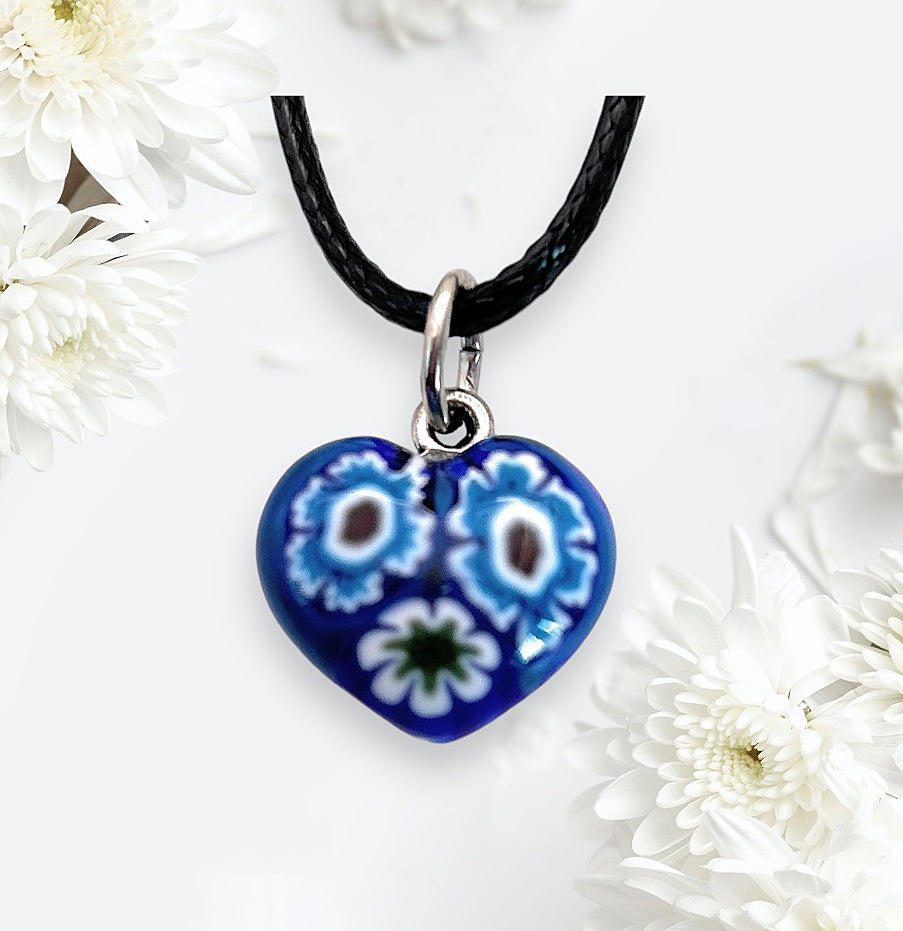 The stunning Murano Glass Pendant by Murano Passion, known as the Murano Heart Pendant - Tiny 1.4cm (MHT1), showcases a blue heart-shaped charm with intricate floral designs, gracefully hanging from a black cord. Encircled by delicate white flowers, it embodies the timeless elegance of Millefiori pieces found in Murano Glass Jewellery.