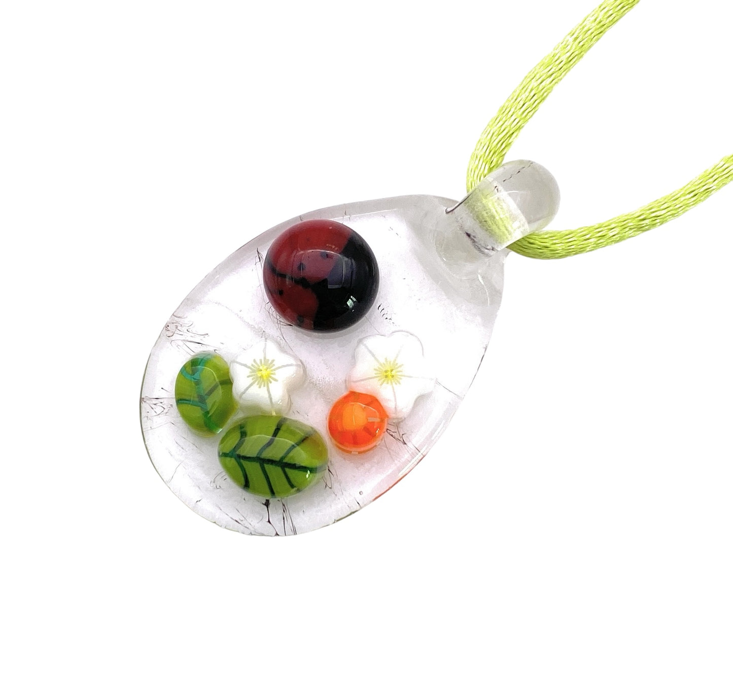 The Calon Glass Ladybird Pendant is a handcrafted glass teardrop adorned with intricate Millefiori designs, featuring a vivid red sphere, a green leaf pattern, and delicate white flowers. Suspended gracefully from a light green cord, this 3cm x 2cm pendant offers an exquisite touch of artistry.