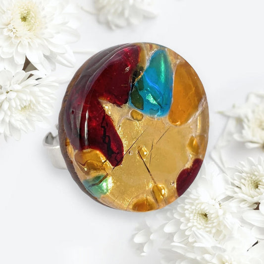 The Murano Glass Ring, a chunky statement piece from Murano Passion, features vibrant abstract shapes in red, gold, teal, and brown. This 3cm genuine ring eloquently embodies Venetian artistic heritage as it rests on a light surface surrounded by delicate white flowers.