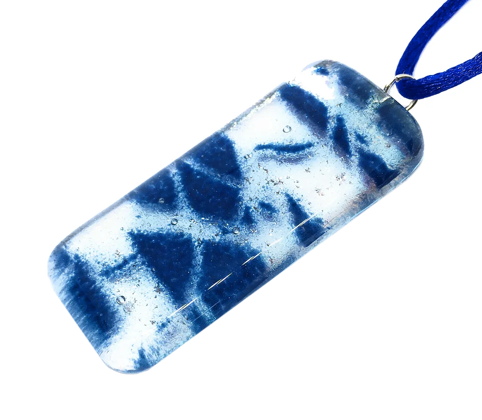 The Dichroic Glass Pendant by Calon Glass features a rectangular design with a smooth, glossy surface that beautifully displays a marbled pattern of blue and white hues, reminiscent of swirling ocean waves. Crafted from handmade glass, this 5cm x 2cm piece is elegantly attached to a blue ribbon for wearing.