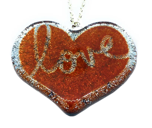 The Handmade 'Love' Pendant in Dichroic Glass by Calon Glass features a heart-shaped design crafted from handmade glass, showcasing a glittery red surface with the word "love" in elegant script. The edges are outlined in sparkling silver, adding a beautiful shimmer. This kiln-fused piece, measuring 4.5cm x 3.5cm, hangs gracefully from a delicate chain, capturing warmth and elegance.