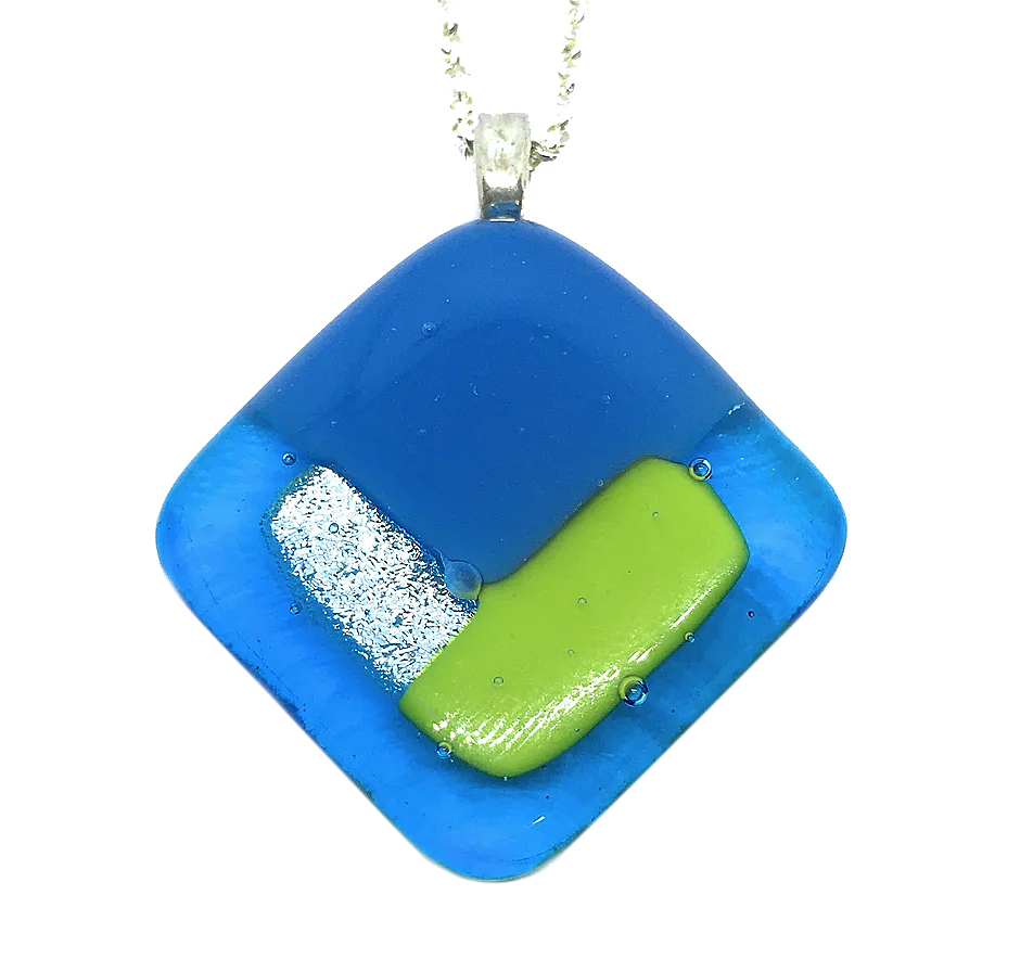 The Handmade Dichroic Glass Pendant by Calon Glass is a stunning 3cm x 3cm square accessory. It showcases a blue background adorned with abstract silver glitter and light green designs. This kiln-fused piece gracefully hangs from a silver chain, providing a unique blend of vibrant artistry.