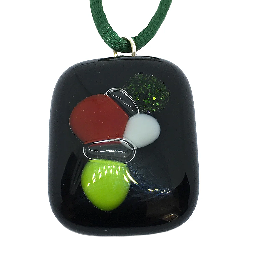 The Handmade Dichroic Glass Pendant by Calon Glass, measuring 3cm x 2.5cm, is presented on a dark green cord and features kiln-fused pieces with a glossy black surface. It is adorned with abstract shapes in red, white, neon green, and glittery dark green details, all meticulously crafted from handmade glass.