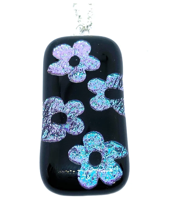 Introducing the Handmade Dichroic Glass Pendant with Flower Design by Calon Glass. Featuring a glossy black background, this exquisite pendant is adorned with five shimmering flowers in delightful shades of pink and blue. Handcrafted from glass, the flowers are strategically spaced to form a beautiful floral pattern and hang gracefully from a silver chain.