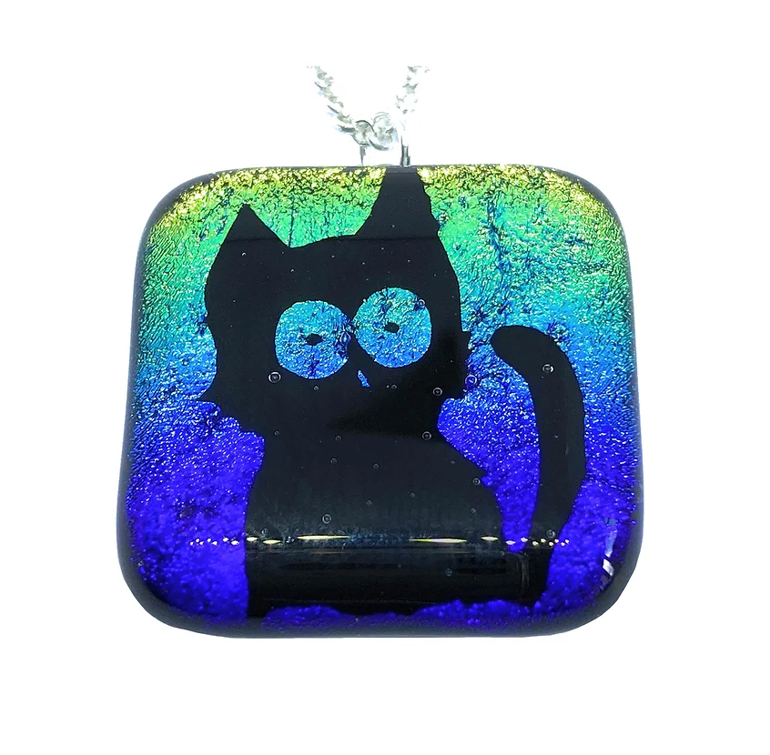 This captivating pendant from Calon Glass showcases a cute cat silhouette on a dazzling dichroic glass background. The handmade design features a mesmerizing gradient that shifts from green to blue to purple, beautifully capturing light. Suspended on a silver chain, this "Handmade Dichroic Glass Pendant with Cute Cat / Lucky Black Cat Design" is truly one-of-a-kind.