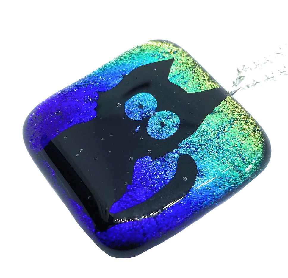 This unique Handmade Dichroic Glass Pendant from Calon Glass features a lucky black cat design with large eyes. The square-shaped pendant, with slightly curved edges, boasts a shimmering background that transitions from green to blue.