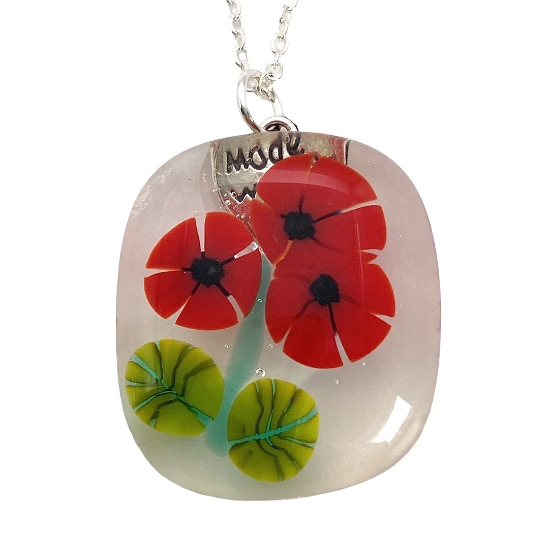 A square-shaped handmade pendant from Calon Glass showcases red poppy flowers and green leaves encased in clear resin, elegantly hanging from a silver chain. The carefully crafted glass piece features a small silver tag with "Made" partially visible at the top.