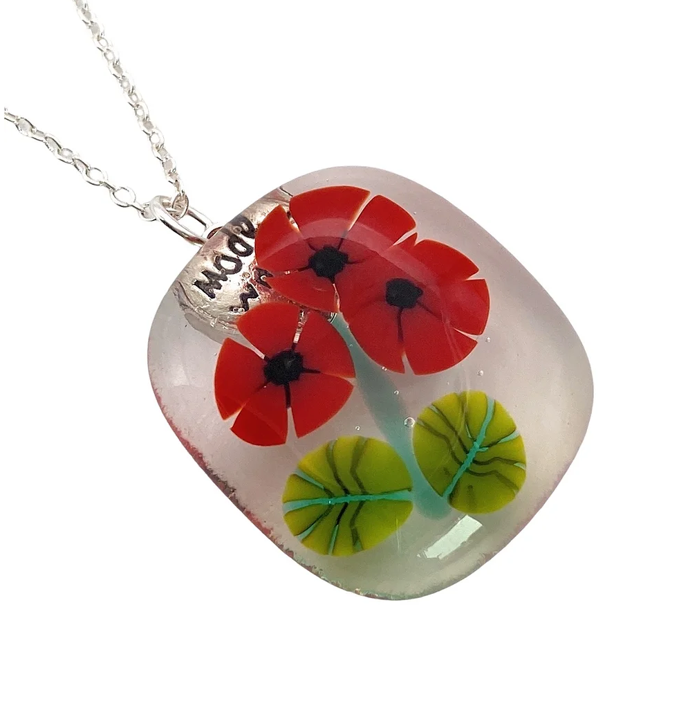 The Handmade Glass Pendant by Calon Glass, also known as the Poppy Pendant or Flower Pendant, showcases vibrant red flowers and green leaves embedded within a square-shaped piece with rounded edges. The pendant comes with a silver chain and features a clear background to highlight the art of kiln-fused glasswork.