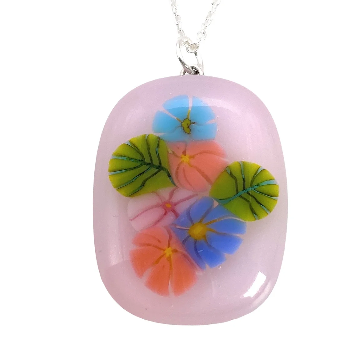 Presenting the Handmade Glass Pendant by Calon Glass: a floral pendant adorned with vibrant blue, pink, and green flowers, artfully set on a smooth oval of handmade dichroic glass. This exquisite piece gracefully hangs from a fine silver chain.