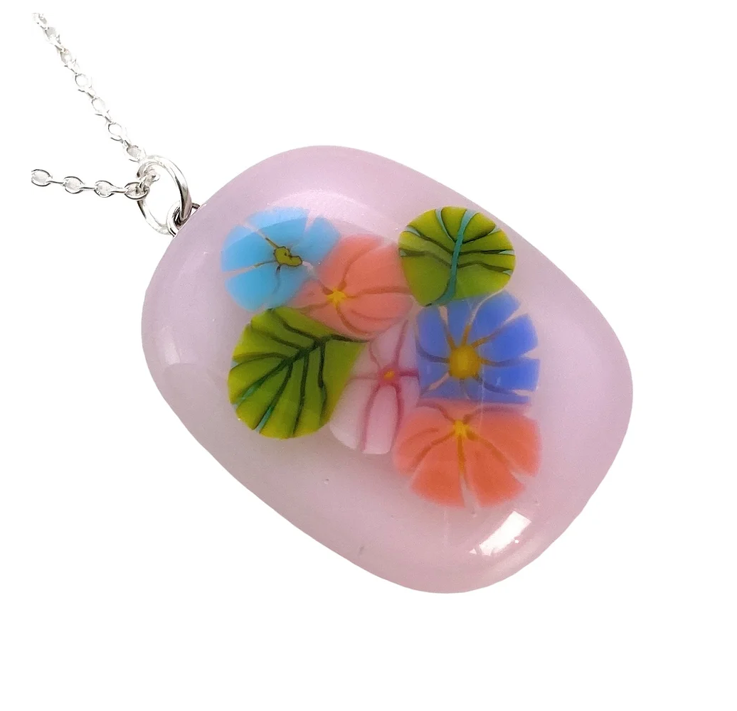 The Calon Glass Handmade Flower Pendant features a pastel pink background adorned with vibrant floral designs in blue, green, pink, orange, and purple. Suspended on a silver chain and crafted from handmade glass, this Floral Pendant captivates with its exquisite charm.