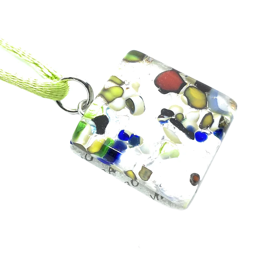 This Murano Passion creation, the Millefiori Pendant on Silver Leaf, is a stunning square piece measuring 2cm x 2cm. It showcases a vibrant mosaic design with scattered patterns of red, blue, green, yellow, and black fragments. The pendant gracefully hangs from a green cord attached to a metal ring.