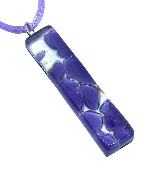 The Murano Glass Pendant on Silver Leaf by Murano Passion features a rectangular design crafted from exquisite purple glass, adorned with a striking mosaic pattern. It hangs elegantly from a delicate purple cord, and its glossy finish highlighted by white accents gives it a vibrant and artistic appearance. This 4cm x 1cm piece of jewelry comes with a Certificate of Authenticity, ensuring its genuine quality.