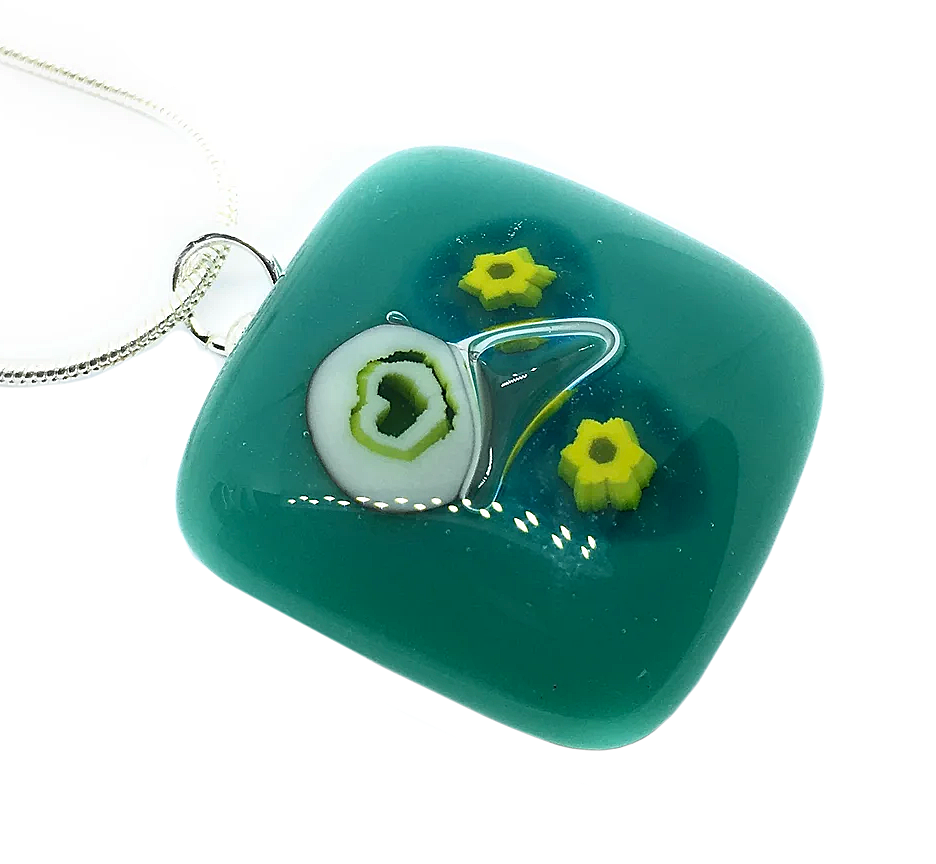 A handmade glass pendant by Calon Glass, featuring millefiori flower designs and a heart, measuring 2.3cm x 2.3cm, with teal glass artistry highlighted in three yellow flowers and a white circle with a green center, complemented by a silver chain.