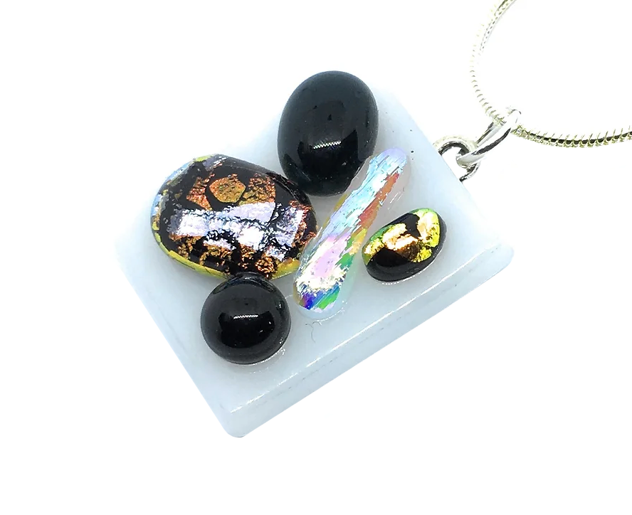 Introducing the Handmade Dichroic Glass Pendant by Calon Glass, a white square pendant measuring 2.3cm x 2cm. It showcases an abstract design adorned with multicolored, shiny stones made from dichroic glass. The stones exhibit vibrant hues of black, gold, and iridescent colors and are elegantly set on a silver chain.