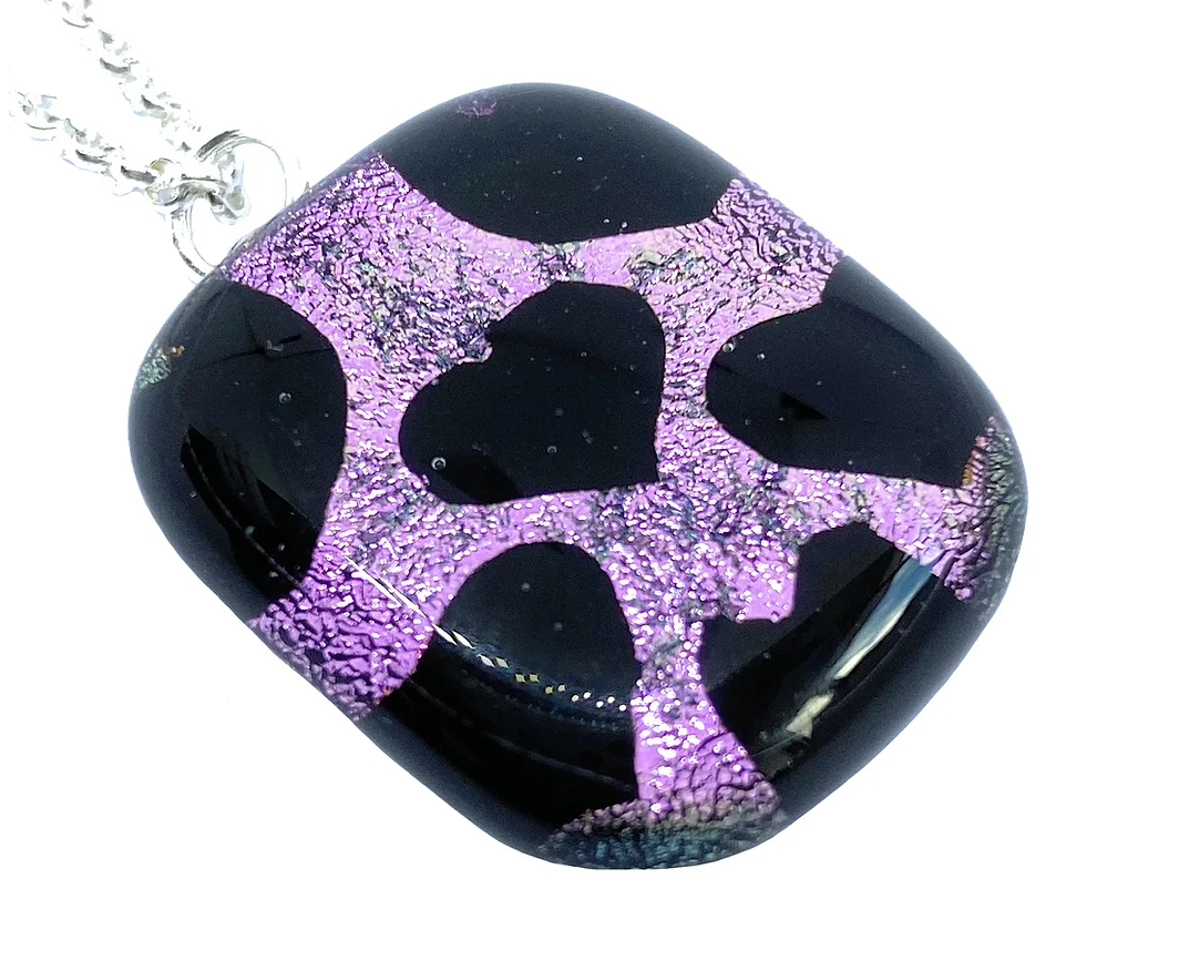 A 2.5cm x 2cm handmade heart-shaped pendant from Calon Glass features shiny purple abstract designs on black dichroic glass and hangs from a silver chain. The intricate design boasts irregular, network-resembling shapes with a textured, reflective surface.