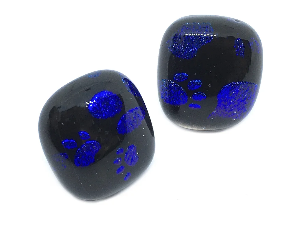 The Handmade Dichroic Glass Earrings by Calon Glass are a pair of eye-catching 1.5cm earrings featuring blue paw print patterns on polished black stones. Set against a white background, these earrings have irregular shapes reminiscent of artisanal glass creations.