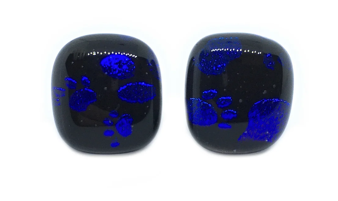 Two black rectangular stones with glossy finishes showcasing sparkling blue paw print patterns are positioned against a white background. These stones, akin to unique artisan jewelry, resemble Calon Glass's Handmade Dichroic Glass Earrings - Paw Print Earrings (1.5cm), featuring rounded edges and a smooth texture.