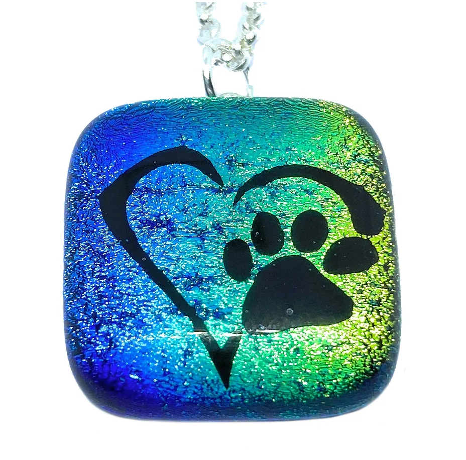 The Handmade Dichroic Glass Pendant by Calon Glass features a shimmering blue and green square, adorned with a black heart and paw print in the center. This elegant piece hangs gracefully from a silver chain and comes beautifully packaged in a gift box, making it an ideal keepsake for animal lovers.