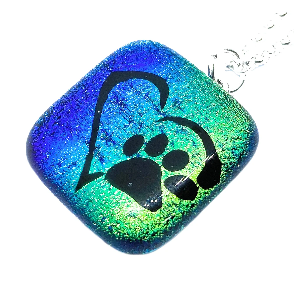 The Handmade Dichroic Glass Pendant by Calon Glass showcases a vibrant blue and green gradient design with a black paw print nestled within a heart outline. This square-shaped piece features a shiny finish and is elegantly suspended from a silver chain, making it an ideal addition to any gift box.