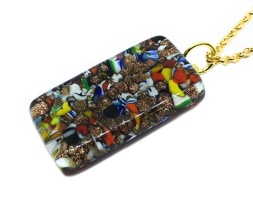 This Murano Passion pendant, named the Millefiori Pendant with Gold Leaf, features a 3cm x 1.5cm rectangular design adorned with a vibrant mosaic of reds, blues, greens, and gold glitter accents. It is elegantly suspended from a gold chain and embodies the alluring craftsmanship of Murano Glass Jewellery. The piece also comes with a Certificate of Authenticity for assurance.