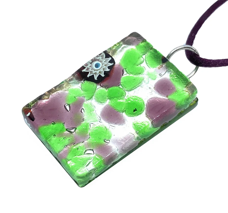 Introducing the Murano Passion's exquisitely crafted rectangular Murano Glass Pendant, featuring a millefiori design in shades of pink and green with a delicate blue and white floral accent at the top. Expertly handmade, this pendant elegantly hangs from a deep purple cord. It comes with a Certificate of Authenticity for added assurance. The dimensions of the pendant are 3cm x 2cm.