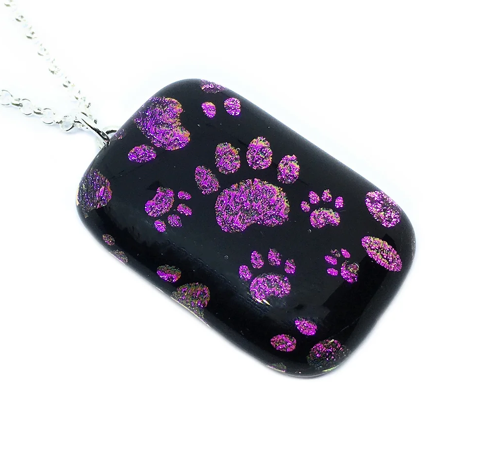 The Handmade Dichroic Glass 'Paw Print' Pendant by Calon Glass showcases a glossy black background adorned with shimmering purple paw prints. Measuring 3cm by 2cm, this distinctive piece hangs gracefully from a silver chain, infusing any outfit with a touch of charm.